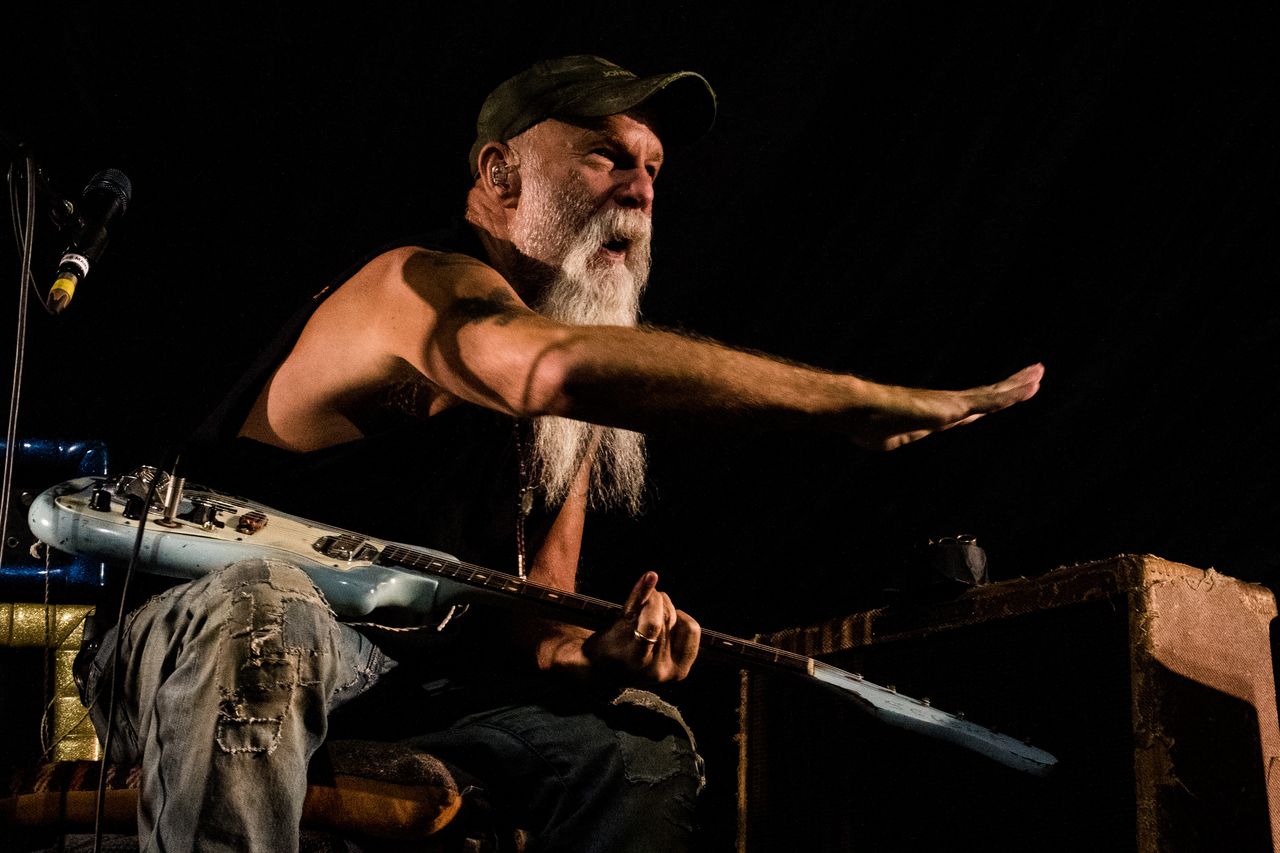 Seasick Steve mit langem weißen Bart sitzt mit Slide-Gitarre auf der Bühne und streckt den Arm aus