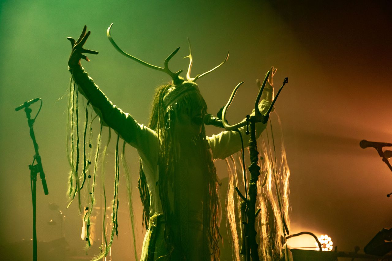 Heilung-Sänger mit Geweih-Kopfschmuck und erhobenen Armen in grün-orangem Licht
